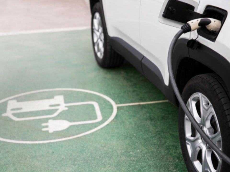 Carro elétrico branco conectado ao cabo de recarga, estacionado sobre símbolo de vaga de carregamento pintado no chão.