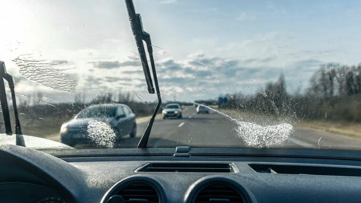Foto tirada de dentro de um carro, que está na estrada, e o seu para-brisa está molhado e as palhetas estão sendo usadas para a limpeza do para-brisa