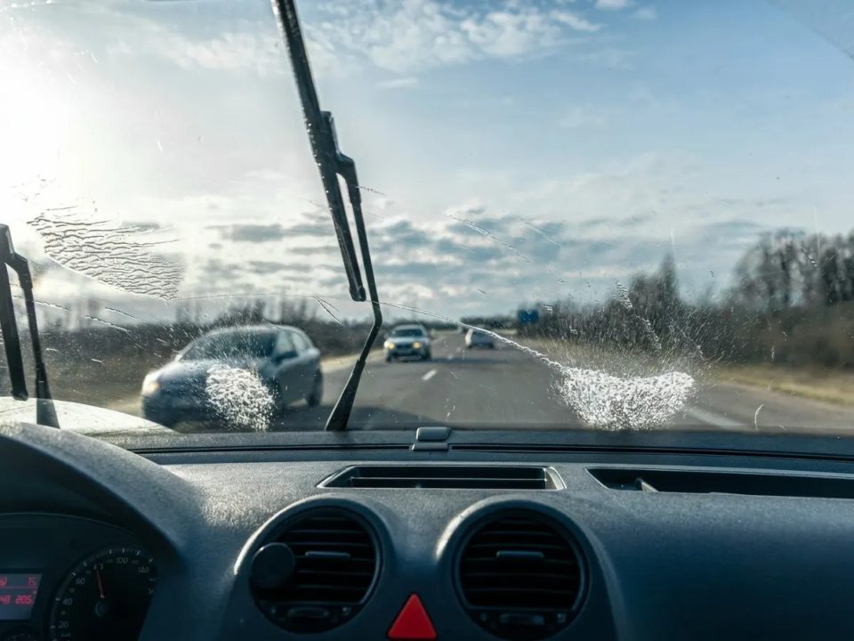 Foto tirada de dentro de um carro, que está na estrada, e o seu para-brisa está molhado e as palhetas estão sendo usadas para a limpeza do para-brisa