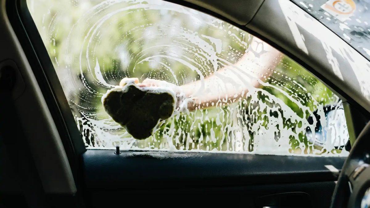 Imagem feita de dentro de um carro, focando no vidro lateral do motorista, que está molhado e com espuma e está sendo esfregado com um pano úmido