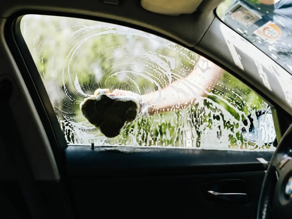 Imagem feita de dentro de um carro, focando no vidro lateral do motorista, que está molhado e com espuma e está sendo esfregado com um pano úmido
