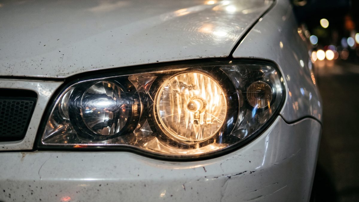 Foto de um farol dianteiro aceso de um carro branco à noite, mostrando a lâmpada halógena iluminada e parte do capô e para-choque