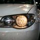 Foto de um farol dianteiro aceso de um carro branco à noite, mostrando a lâmpada halógena iluminada e parte do capô e para-choque