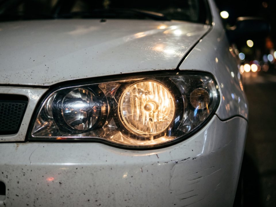 Foto de um farol dianteiro aceso de um carro branco à noite, mostrando a lâmpada halógena iluminada e parte do capô e para-choque