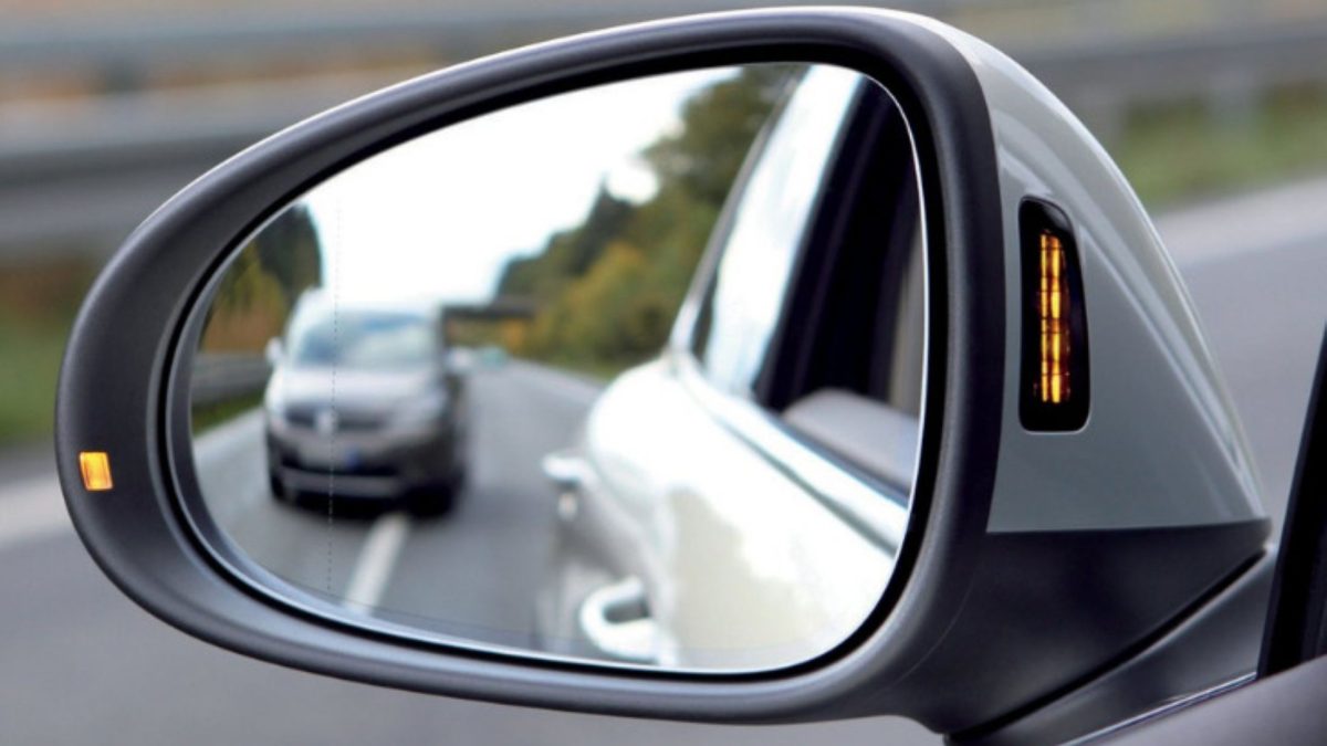 Retrovisor lateral de carro com luz indicadora de ponto cego acesa, refletindo outro veículo na estrada.