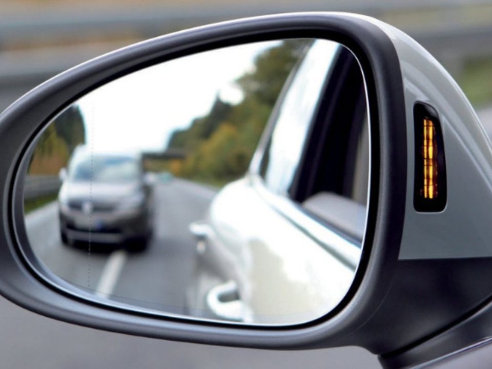 Retrovisor lateral de carro com luz indicadora de ponto cego acesa, refletindo outro veículo na estrada.