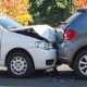 Colisão traseira entre um carro branco e um carro cinza, com o para-choque dianteiro amassado.